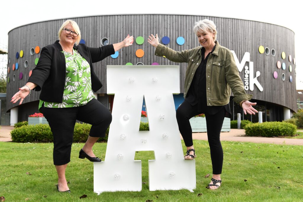 Maria Bowness (left) and Sally Alexander (right) from Milton Keynes College Group are stood outside the MK College Chaffron Way building, with one foot each on a large letter 'A'.
