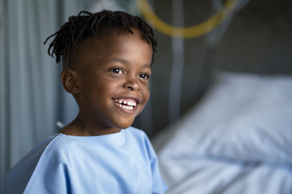 Smiling boy looking away sitting on bed at ward. Cute male patient is in blue gown. He is at hospital.