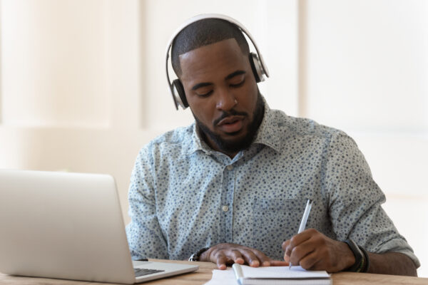 Focused student sit at desk wearing wireless headphones learning preparing for seminar or exam, guy interpreter hears audio writing down translation, online lecture course e-learning concept