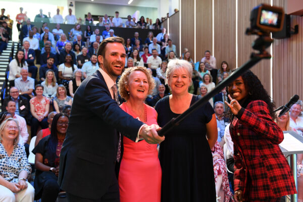 Alex Warner from MK College Group holds a selfie stick and takes a photo with the three other speakers at the SCIoT launch event with a crowd of people behind them.