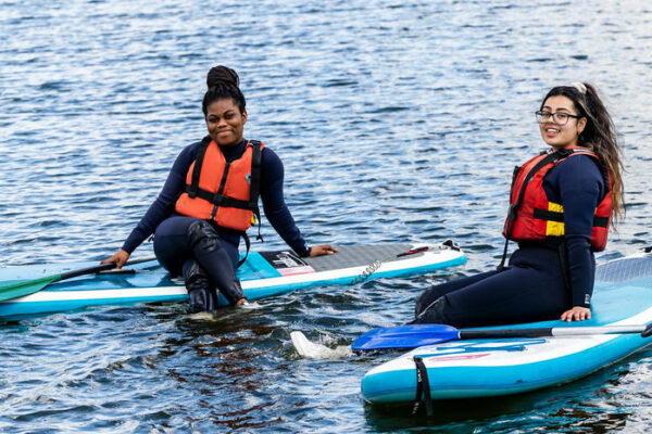 2 female public services students in the middle of the lake sat on paddle boards.
