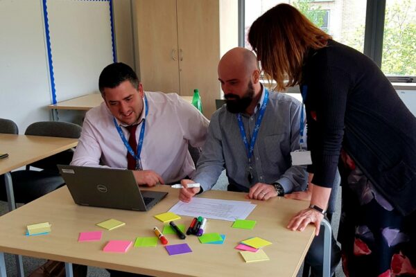 3 teachers sat at a desk looking at a laptop while making notes on coloured post-it stickers.