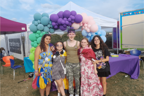 A group of five people all wearing bright colours. They are standing in a line close together outside in front of a rainbow coloured balloon arch.