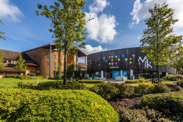 The front of the building of MK College's Chaffron Way campus. The building is clad in dark wood covered with colourful spots. In the foreground are green trees and bushes.