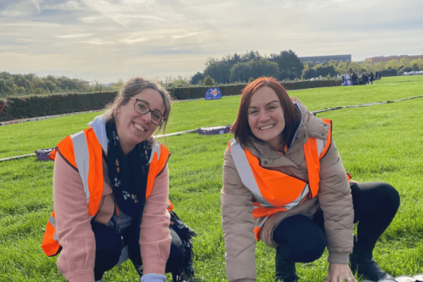 Two colleagues from Milton Keynes College Group at MK Can event. They are crouched down in an open field with rows of canned food behind them, wearing winter clothes and orange hi-vis vests.
