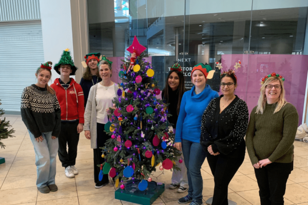 Staff and students from Milton Keynes College are stood in a line, wearing Christmas jumpers and Christmas headbands. In the middle is a Christmas tree covered in pink, yellow and blue Christmas baubles.