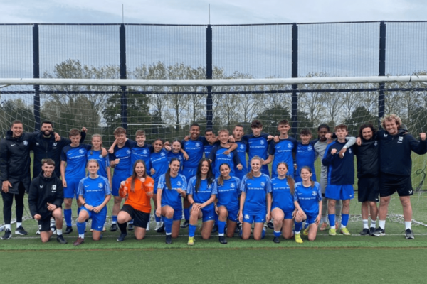 MK College Women's and Men's Football Academy teams, wearing blue football kits. The teams are standing in the centre of a football goal with their coaches standing at the sides.