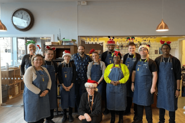 Students and staff from MK College are stood in front of the coffee counter at the College's Brasserie restaurant. Everyone is stood in two lines and they're all wearing blue aprons and red Santa hats.
