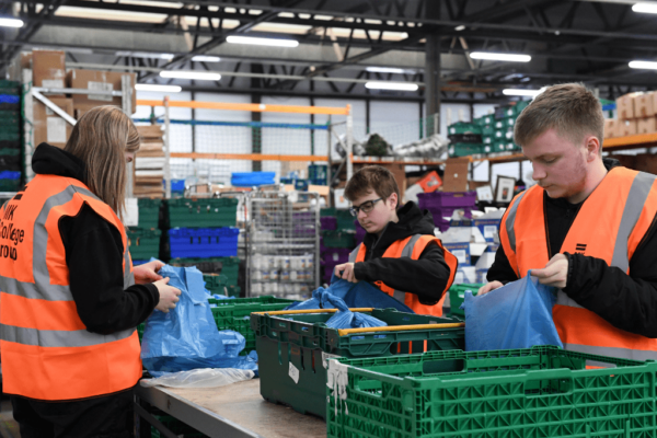 Three MK College students in orange high vis vests in a warehouse packing food into crates.