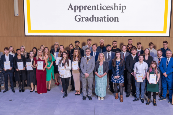 Apprentices from MK College Group at their graduation ceremony with the Mayor and Mayoress of Milton Keynes in the centre of the front row. The apprentices are stood in front of a screen that says 'Apprenticeship Graduation'.