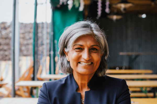 Chair of Governors at MK College Group, Ruby Parmar. Ruby is sat down, directly facing the camera and smiling, wearing a dark blue suit with a green and blue background.