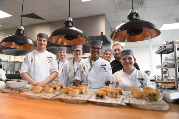 Commis Chef appretnices from MK College are in their chefs' whites standing in the kitchen at the College's Brasserie restaurant. At the table in front of them are trays of cakes that have been prepared as part of a community lunch for the College's College in the Community Day.