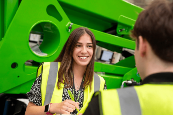 MK College apprentice Evie Savage is in a yellow high vis jacket in front of a large green machine at her employer Niftylift's headquarters.