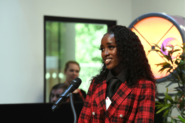 Former SCIoT student Deborah Mungai addresses the audience at the SCIoT opening event. Deborah is smiling and is wearing a red and black checked suit.