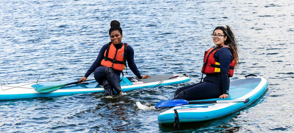 2 female public services students in the middle of the lake sat on paddle boards.