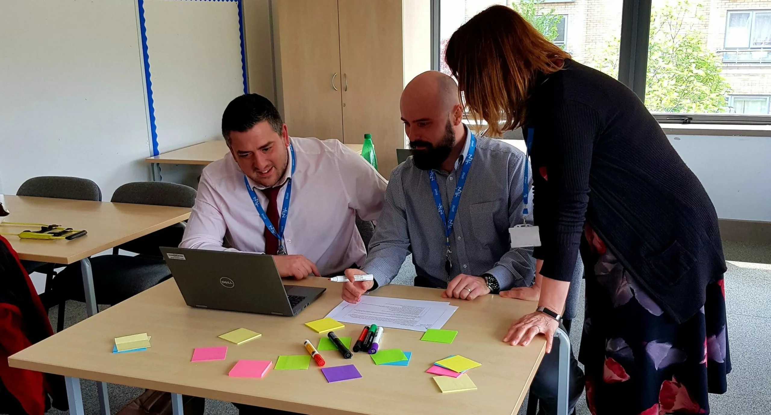 3 teachers sat at a desk looking at a laptop while making notes on coloured post-it stickers.