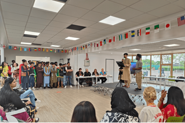 Students and staff from Milton Keynes College celebrating the College's Cultural Diversity Week. There are two students standing on a stage addressing an audience of staff and students - some people are standing, some are sitting and many are wearing national dress.