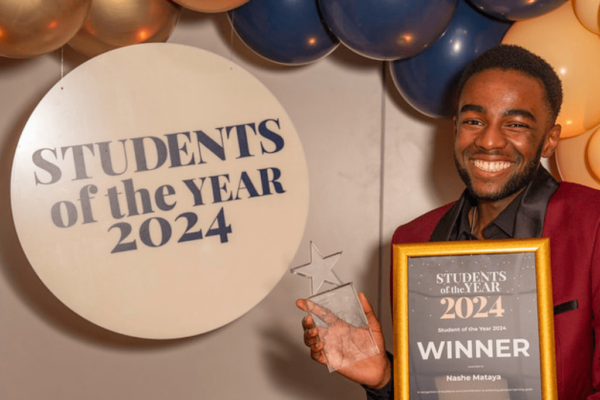 Nashe Mataya, winner of the overall Milton Keynes College Group's Student of the Year Award. Nashe is standing next to a round sign that says 'Students of the Year 2024' and is holding a framed certificate and trophy. Nashe is smiling and wearing a red suit jacket.