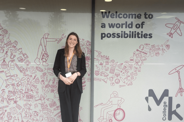 Lauren Gallyot - the new Sustainability Lead for Milton Keynes College Group - is standing outside of the Chaffron Way campus, in front of a door with a mural with the words 'welcome to a world of possibilities' and the MK College logo.