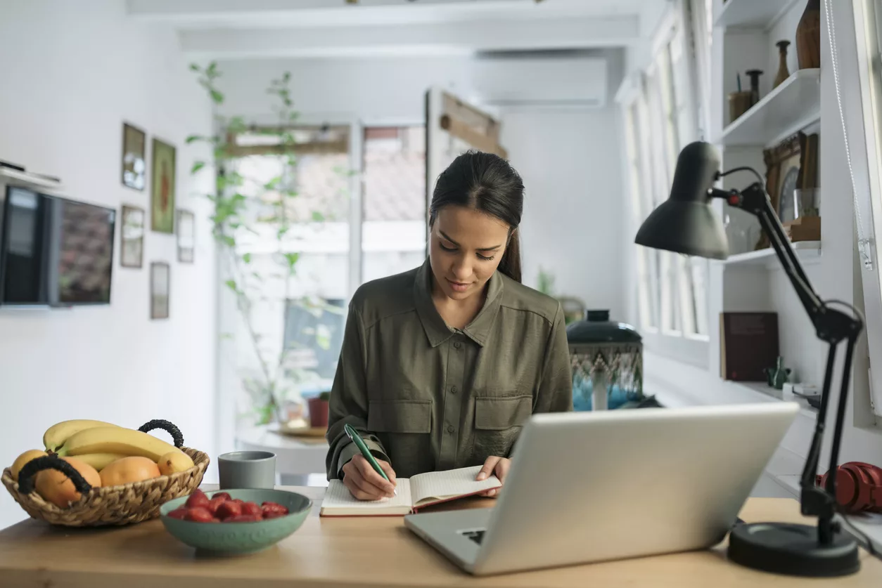 Unemployed beautiful young woman looking for a job online from home using laptop and writing down the notes in her notebook