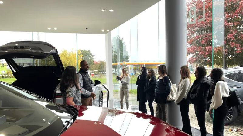 Students from MK College at a Steven Eagell car dealership. The students are stood next to a red car while a staff member is talking to them.