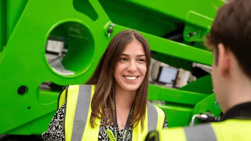 College student wearing a green vest, a lanyard and holding a pen. She is speaking to another student and smiling. On the background, there is green machinery.