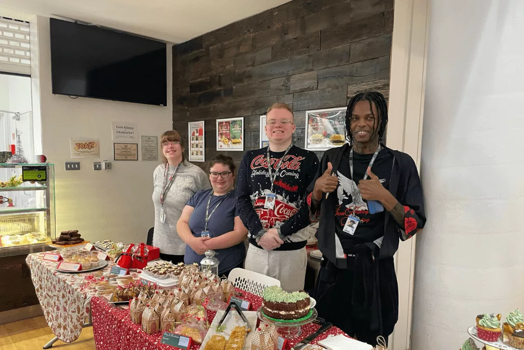 Inclusive Learning students from MK College are stood in a row in front of a table of cakes.