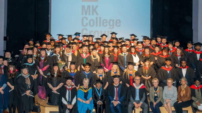 STudents at MK College Group's HE Graduation at the Stables Theatre. The students are on the stage, wearing graduation gowns and hats.