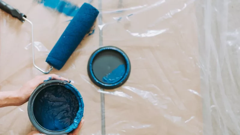 A photograph taken from above of hands holding an open tub of blue paint. The lid of the tub and a used paint roller lie on the floor on top of a semi-transparent plastic cover.