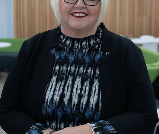 Maria Bowness, Group Director for Curriculum and Pedagogy at MK College Group. Maria is leaning against a desk and looking straight at the camera.