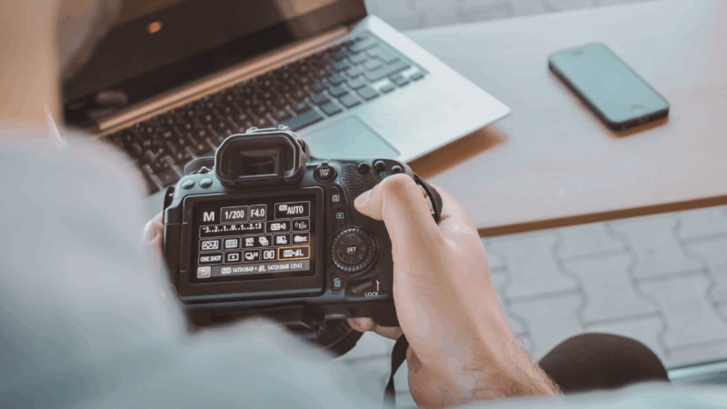 A person holding a DSLR camera, adjusting settings on the screen while sitting at a desk with a laptop and phone.