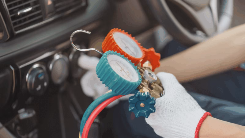 A person wearing gloves holding refrigerant pressure gauges inside a car with dashboard controls in the background.