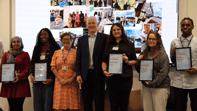 MK College students at the HNBC internship celebration event. The students are stood in a line holding certificates, with a powerpoint presentation behind them showing photos from during their internships.