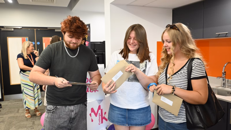 Students from MK College opening their A Level results. Three students stood in a line opening their envelopes with their results in.
